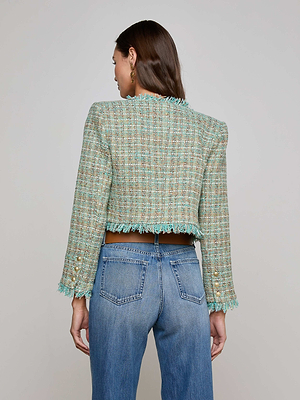 A woman with long brown hair wears blue jeans and the L'AGENCE Sale Clydie Cropped Tweed Jacket, featuring a collarless neckline, fringe details, and gold-tone sleeve buttons. She stands with her back to the camera against a plain background.