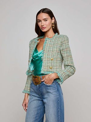 A woman poses in the L'AGENCE Sale Clydie Cropped Tweed Jacket, a pastel green collarless blazer with gold-tone buttons, over a shiny green satin top, styled with blue jeans, a wide brown belt, long brown hair, and gold earrings.