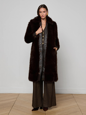 A woman with straight dark hair and gold earrings stands indoors against a light wall, wearing the L'AGENCE Irisa Faux Fur Coat in chocolate over a gray buttoned dress, brown wide-leg pants, and brown pointed shoes.