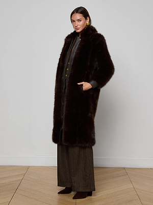 A woman stands against a plain white wall, wearing the L'AGENCE Irisa Faux Fur Coat in chocolate over a dark outfit with wide-leg trousers and heeled boots. She has one hand in her pocket and looks at the camera.
