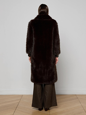 A person with straight, dark hair faces a plain wall, wearing the L'AGENCE Irisa Faux Fur Coat in rich chocolate and wide-leg pants. The soft-textured coat extends just below the knees and is viewed from behind.