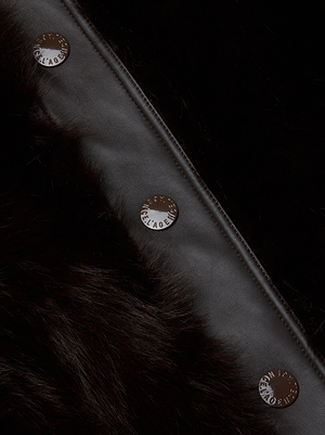 A close-up of the Irisa Faux Fur Coat by L'AGENCE in chocolate, featuring three round metal buttons engraved with the brand name, aligned vertically along a leather trim.