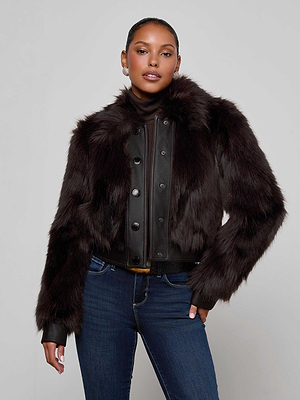 A woman with long hair models the Claira Faux Fur Jacket by L'AGENCE—featuring leather trim—layered over a black turtleneck and blue jeans, standing confidently against a light gray background to showcase this cold weather essential.