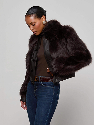 A woman models the Claira Faux Fur Jacket by L'AGENCE—featuring dark, long hair faux fur and leather trim—styled with a brown turtleneck, blue jeans, and gold earrings against a plain background. A true cold weather essential.