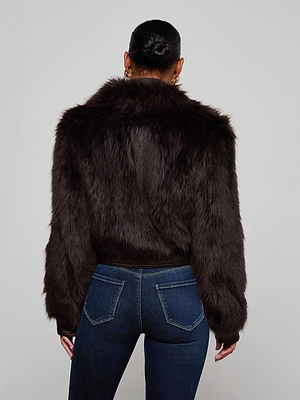 A woman stands with her back to the camera in a Claira Faux Fur Jacket by L'AGENCE, featuring dark brown faux fur with leather trim, styled with blue jeans and hoop earrings. Her hair is in a neat bun, set against a light background.