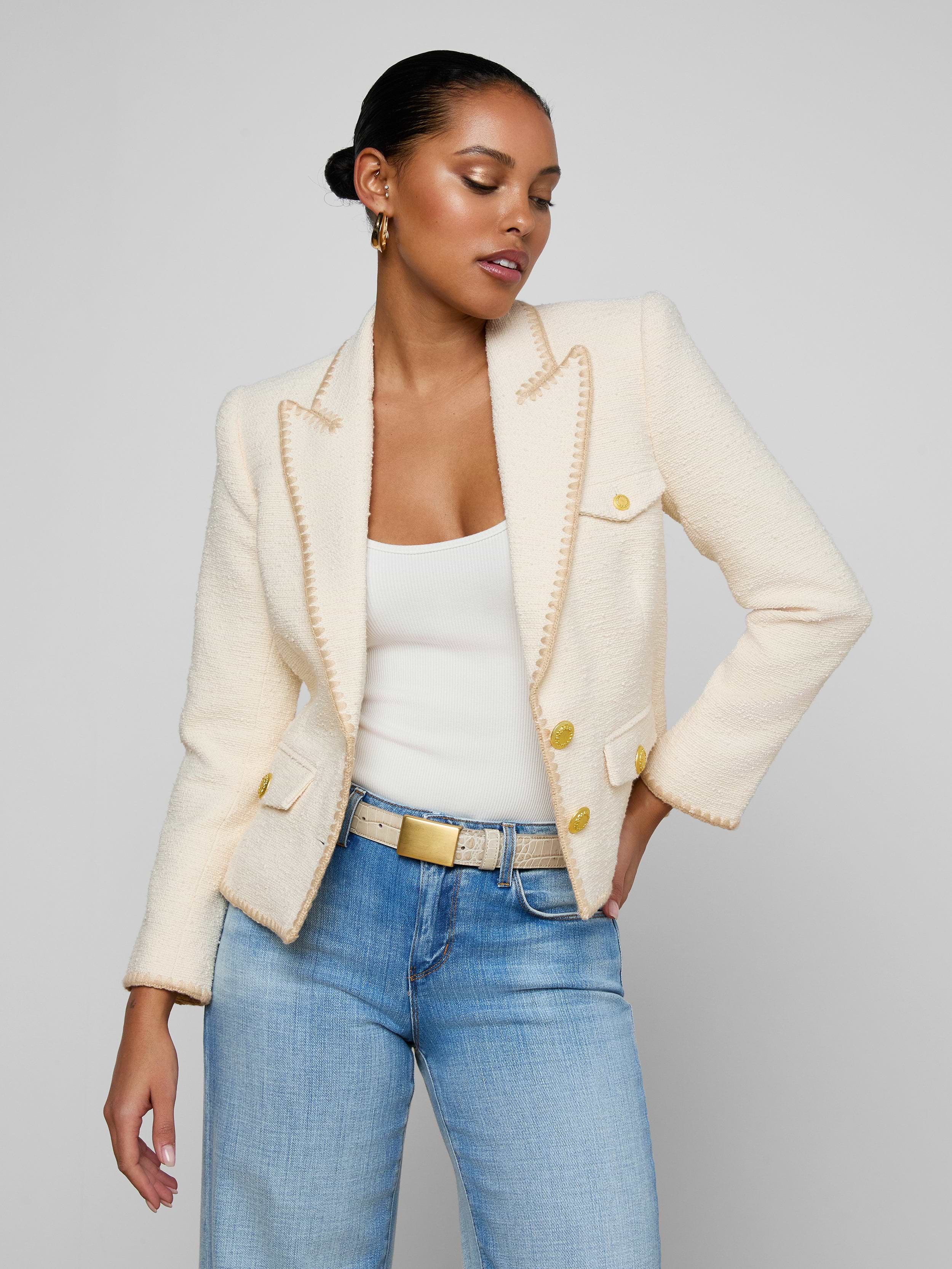 A woman stands against a plain background with one hand in her pocket, wearing the Wylla Tweed Blazer in neutral ecru with gold buttons, paired with a white top and light blue jeans.