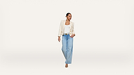 A woman stands against a plain white background, wearing a cream blazer, white top, light blue wide-leg jeans, and sandals. She looks to her left with one hand on her blazer and one relaxed by her side.