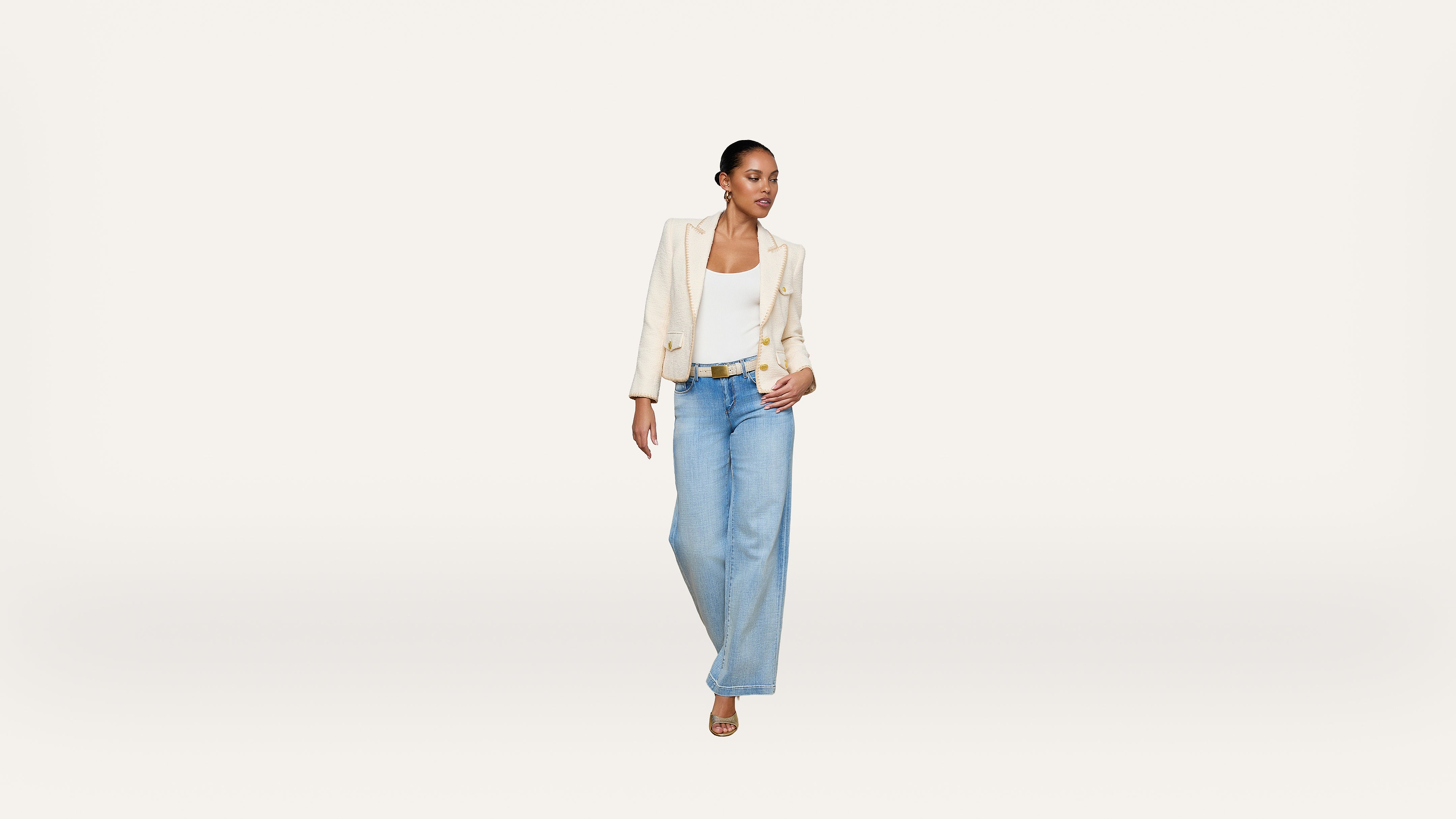 A woman stands against a plain white background, wearing a cream blazer, white top, light blue wide-leg jeans, and sandals. She looks to her left with one hand on her blazer and one relaxed by her side.
