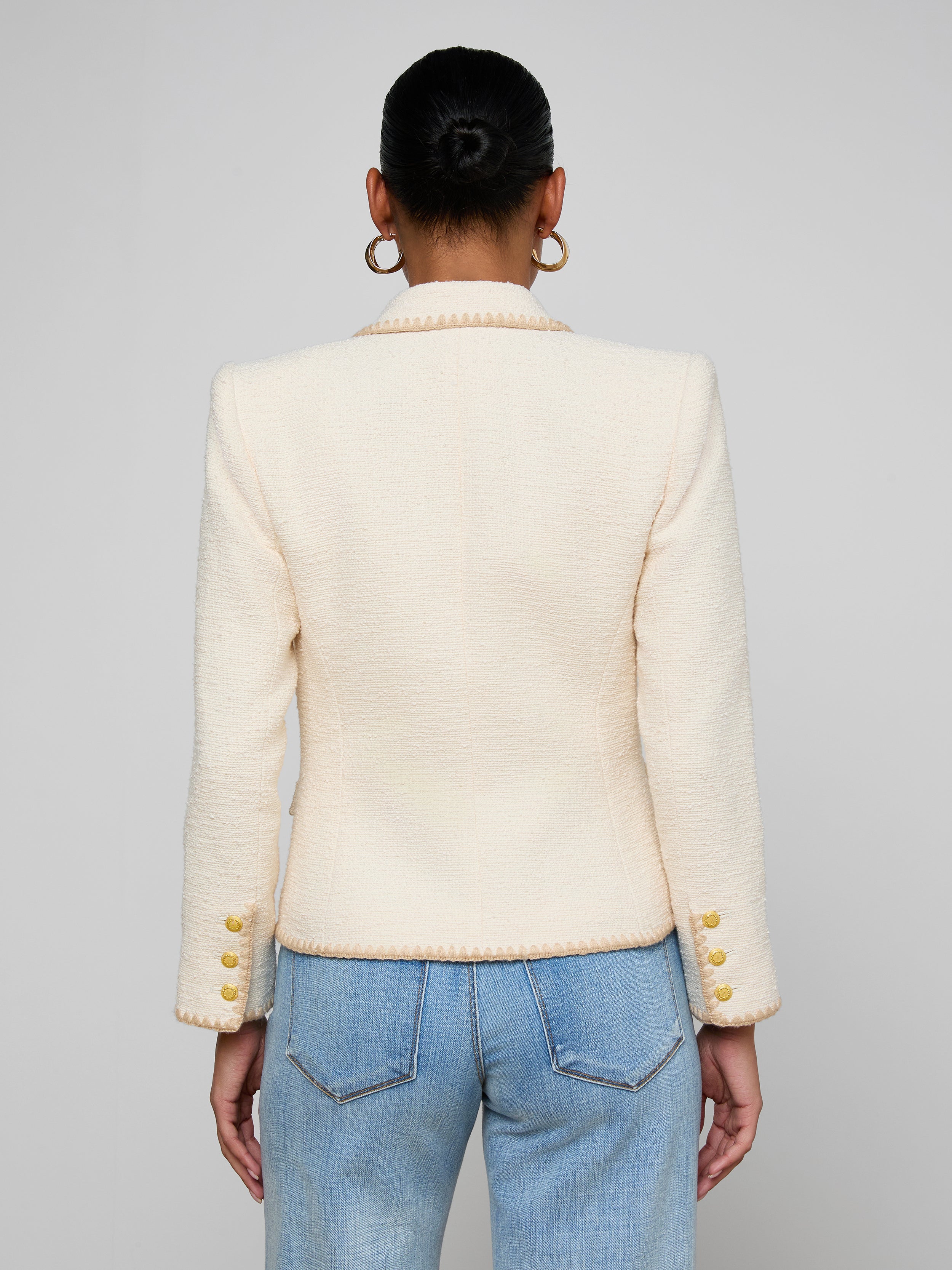 A person with dark hair in a bun wears the Wylla Tweed Blazer in a neutral ecru shade, featuring gold buttons, paired with light blue jeans. They are facing away from the camera against a plain off-white background.