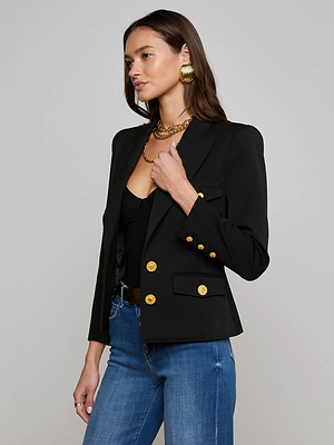 A woman wearing the Wylla Blazer with gold buttons, a black top, blue jeans, large gold earrings, and a gold necklace stands against a plain light gray background.