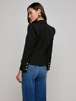 A woman with long dark hair faces away, wearing the Wylla Blazer with gold buttons and wide-leg blue jeans. She pairs it with large gold hoop earrings against a plain light background.