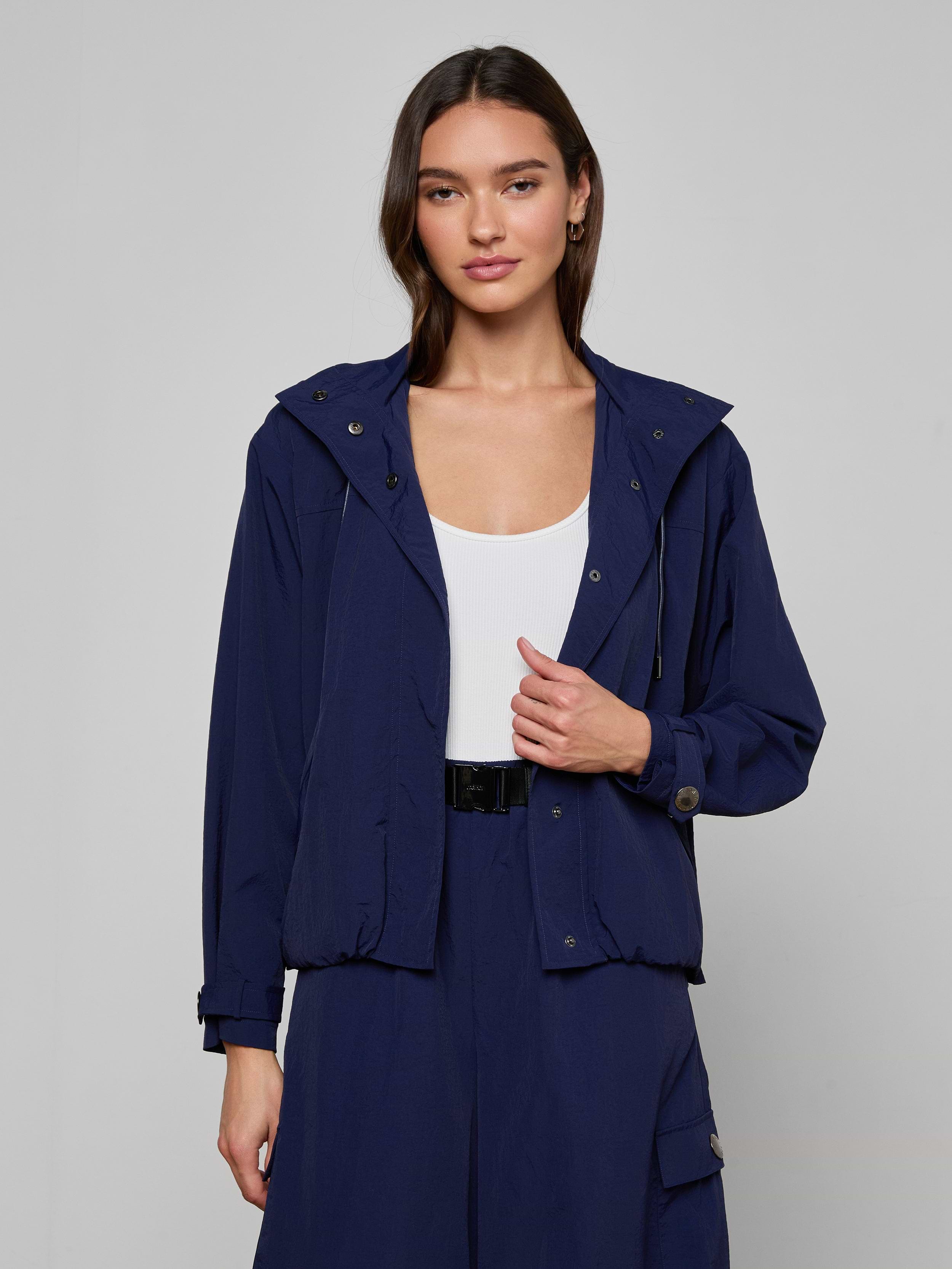 Wearing the Geovana Hooded Windbreaker in navy blue over a white top and matching navy pants, a woman with long brown hair stands against a plain light background.