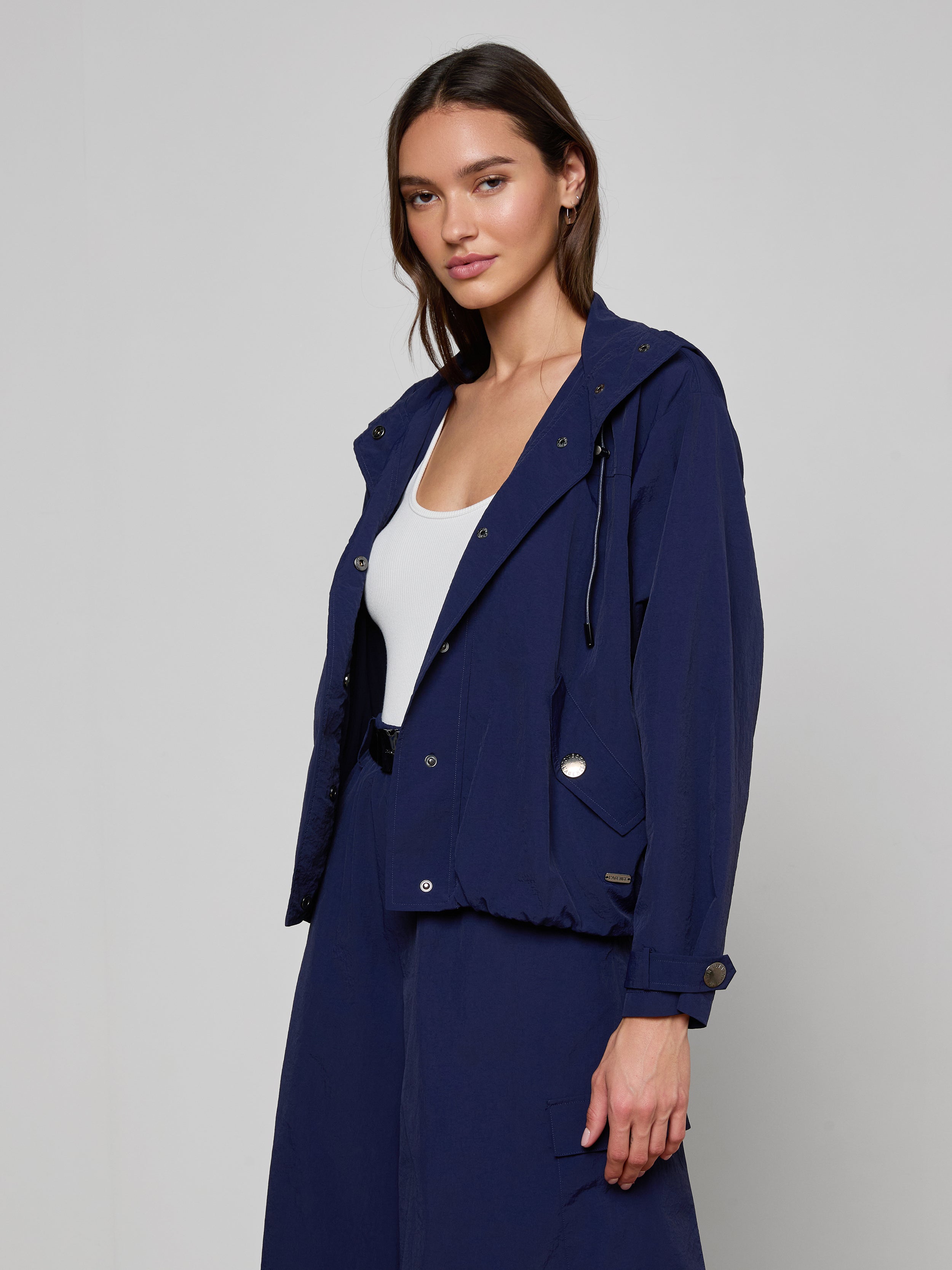 A woman wearing the Geovana Hooded Windbreaker in navy blue over a white top stands against a plain light background, looking slightly to the side with a relaxed expression.