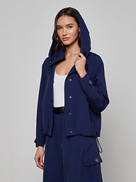 Wearing the Geovana Hooded Windbreaker in navy blue, which features a removable hood, over a white top and matching navy blue pants, a woman stands against a plain light background.