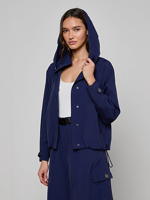 A woman faces forward in a plain, light-colored setting, wearing the Geovana Hooded Windbreaker in navy blue with matching pants over a white top, holding her hood. This water-repellent jacket is an ideal travel essential for any weather.