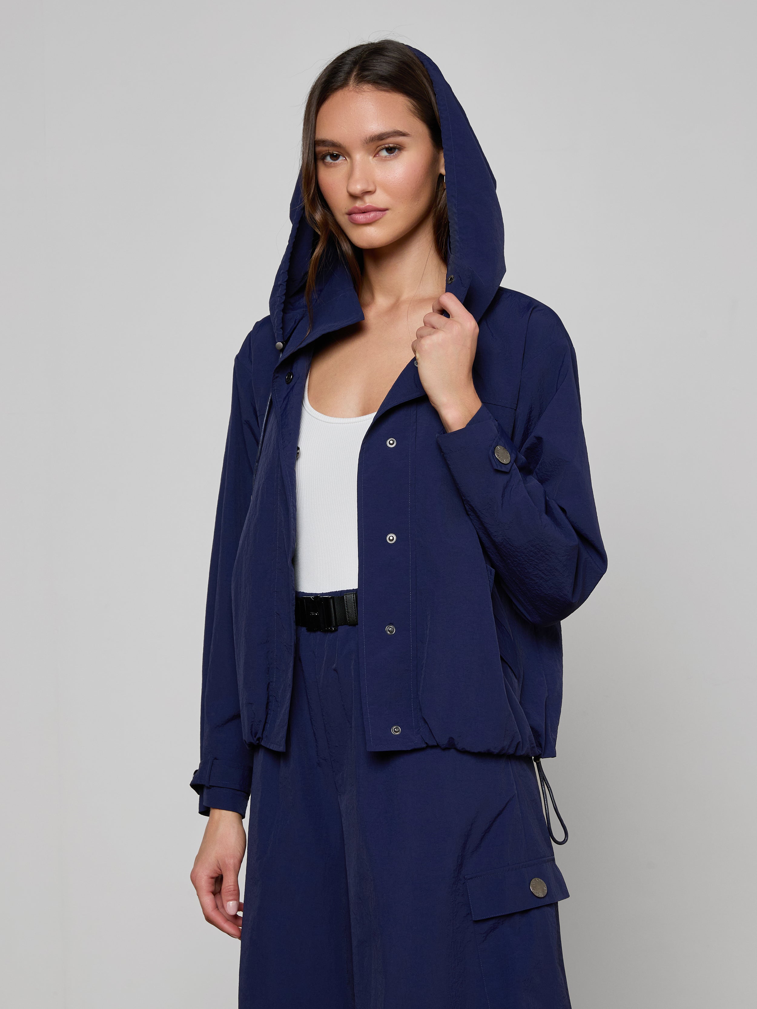 Wearing the Geovana Hooded Windbreaker in navy blue, which features a removable hood, over a white top and matching navy blue pants, a woman stands against a plain light background.