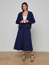 A woman wearing the Geovana Hooded Windbreaker in navy blue, paired with a matching midi skirt, a white top, and black strappy heels, stands against a plain white background.