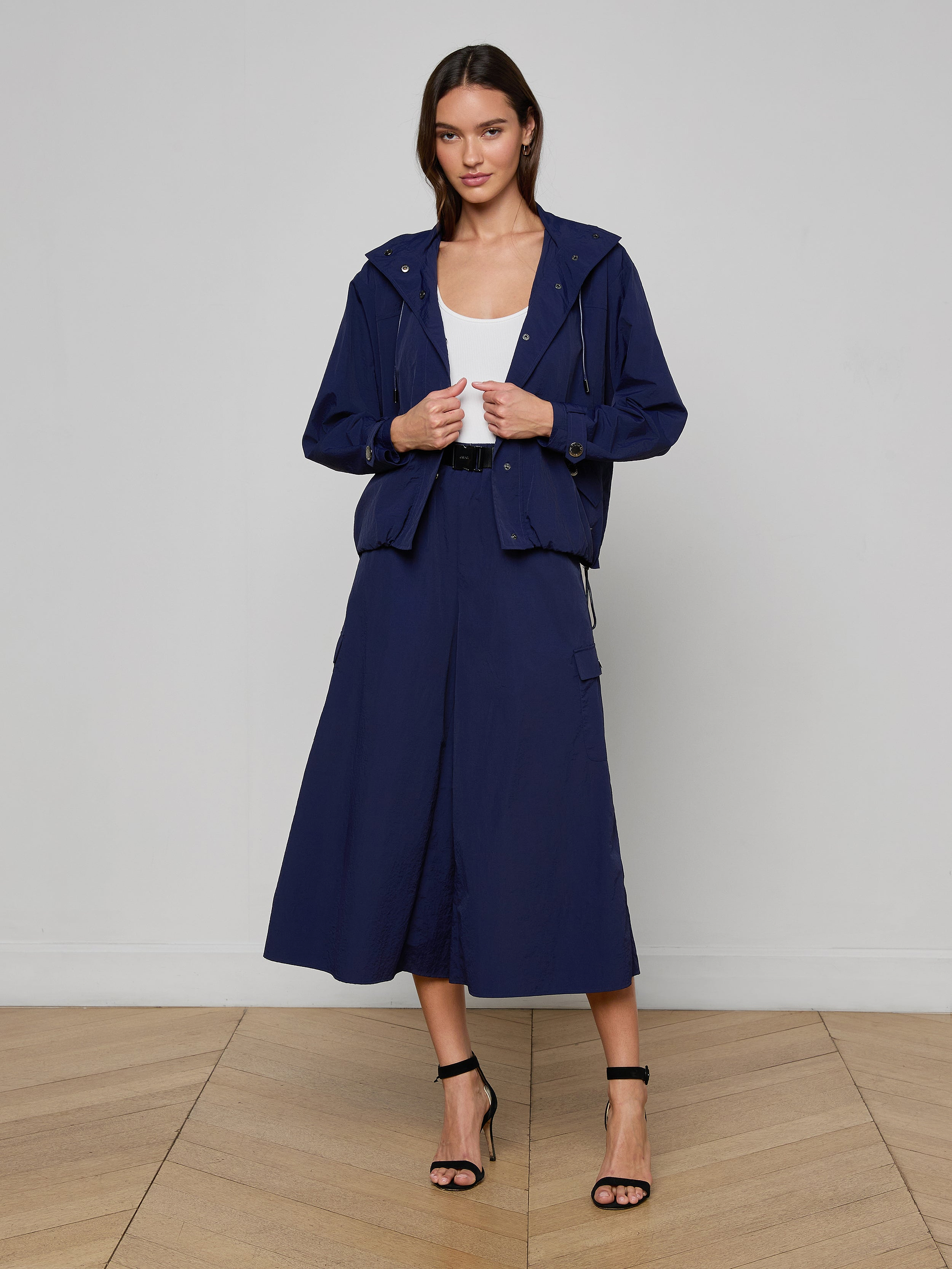 A woman wearing the Geovana Hooded Windbreaker in navy blue, paired with a matching midi skirt, a white top, and black strappy heels, stands against a plain white background.