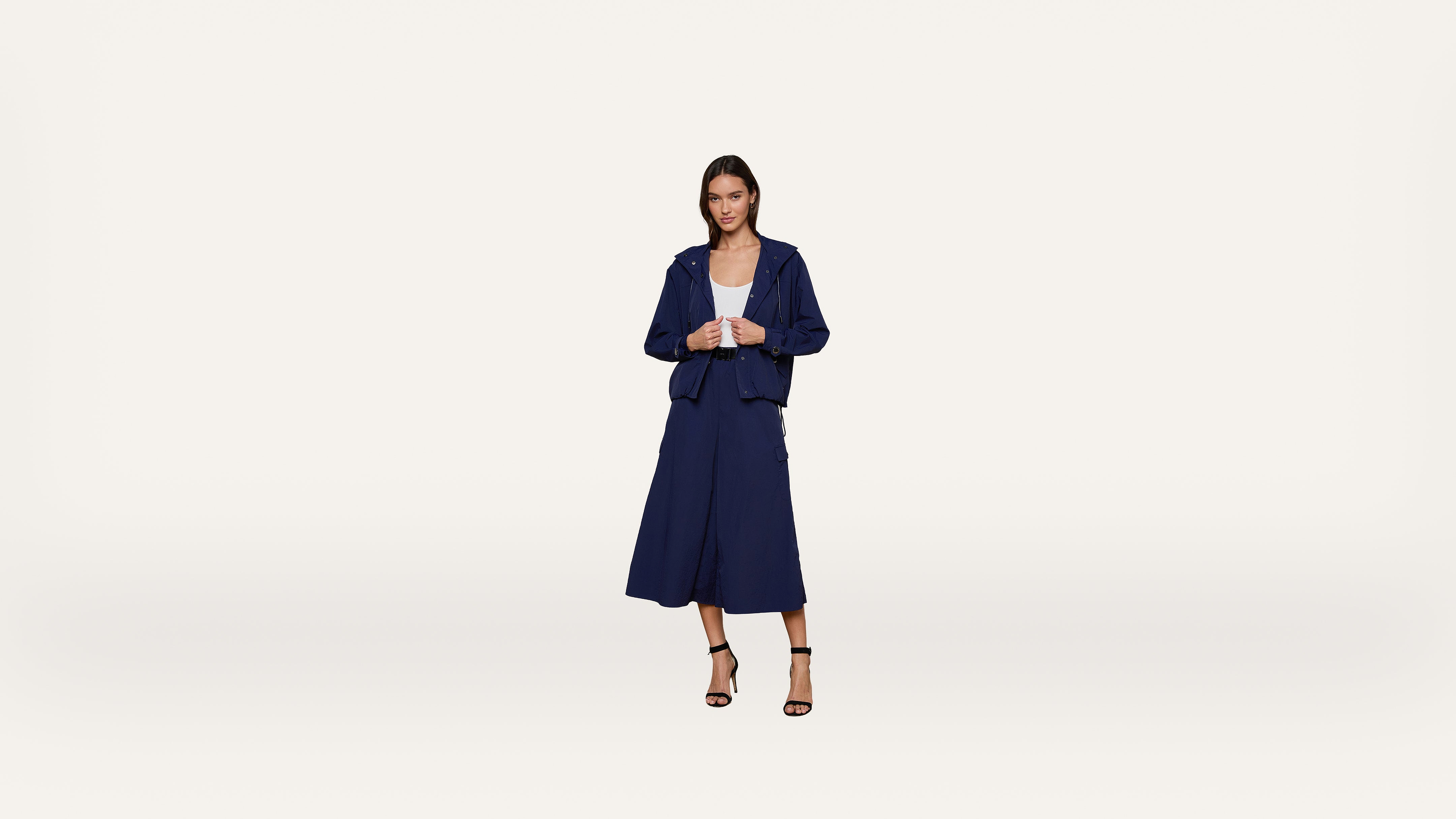 A woman stands against a plain white background wearing a navy blue jacket and culottes, a white top, and black heeled sandals.