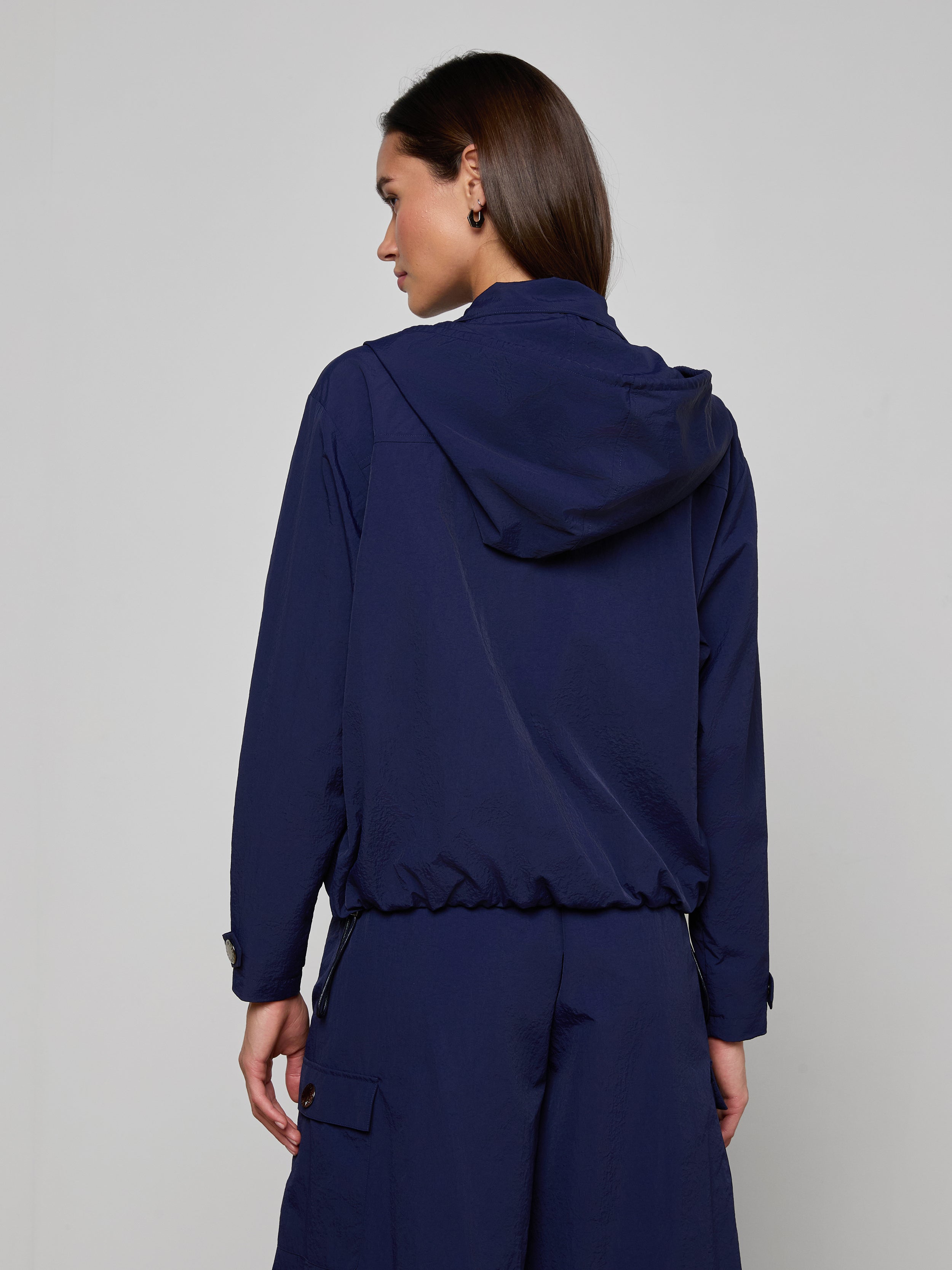 Facing away from the camera, a woman wears the Geovana Hooded Windbreaker in navy blue with matching pants, standing against a plain light background. Her dark hair is tucked behind her ear, and her left arm hangs relaxed at her side.