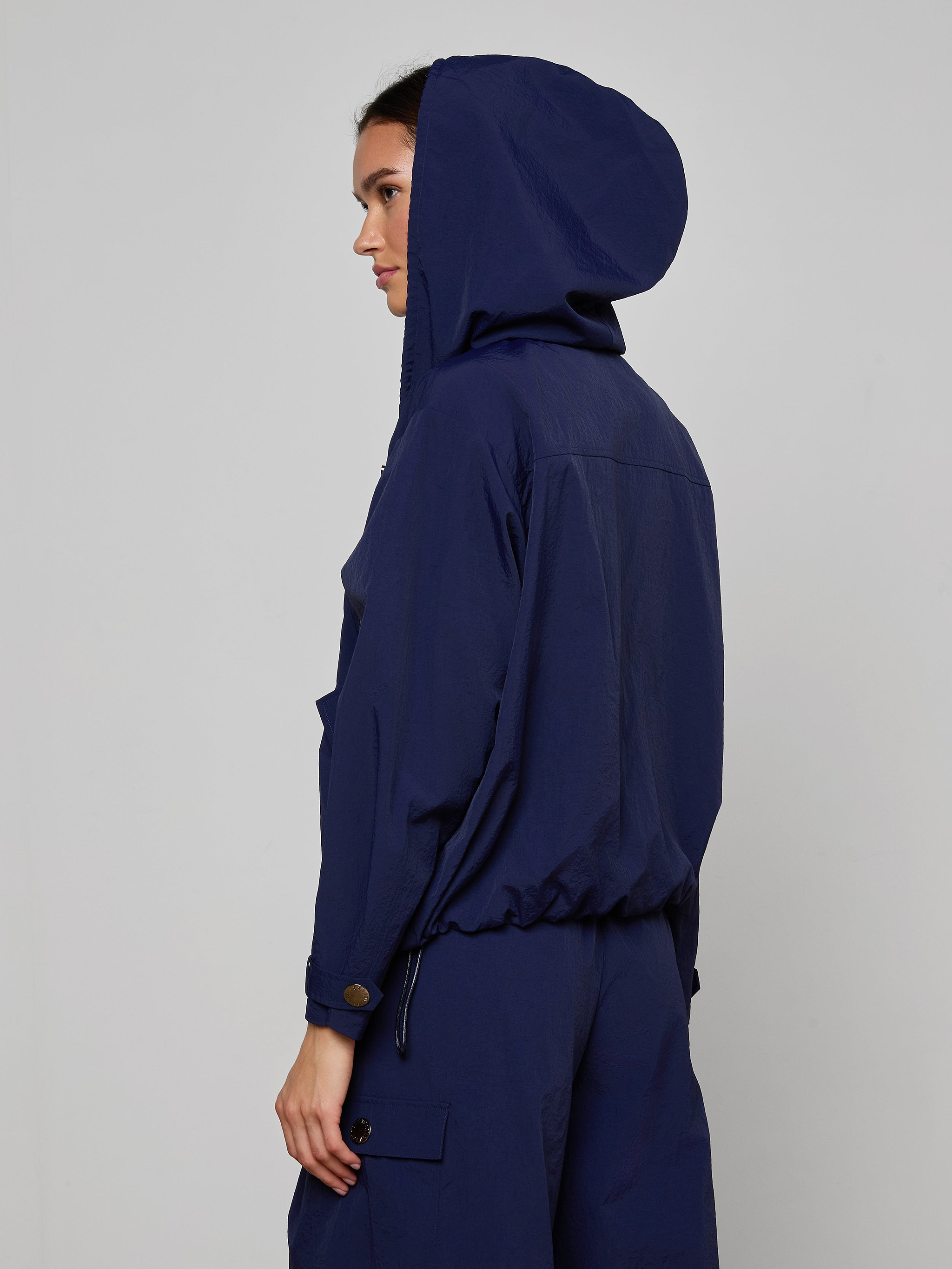 A person stands in profile against a plain, light background, wearing the Geovana Hooded Windbreaker—a navy blue water-repellent jacket—paired with matching pants.