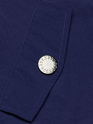 A close-up of the Geovana Hooded Windbreaker’s blue, water-repellent fabric with a silver button stamped LAGENCE. Visible stitching runs diagonally beside the button.