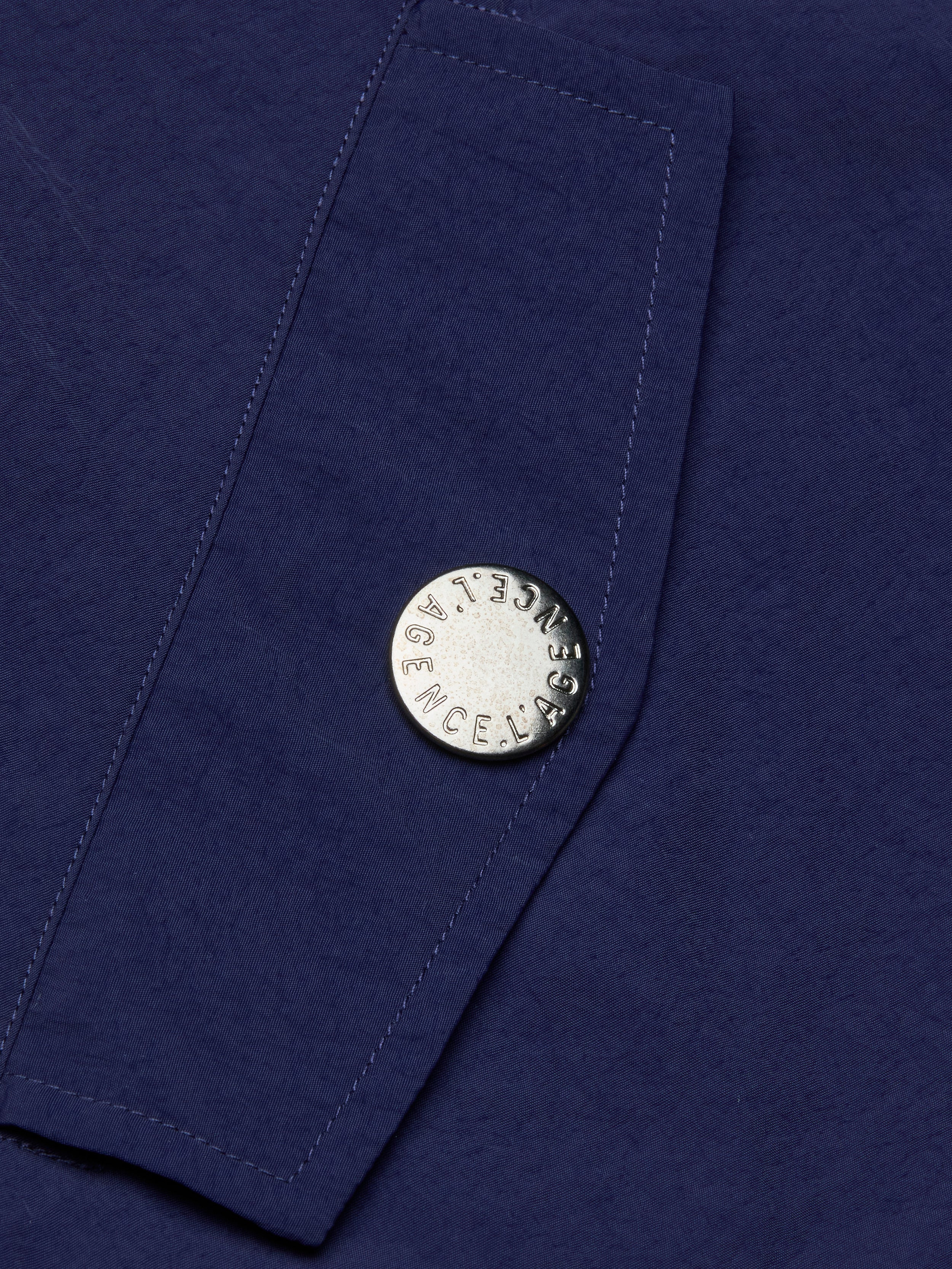 A close-up of the Geovana Hooded Windbreaker’s blue, water-repellent fabric with a silver button stamped LAGENCE. Visible stitching runs diagonally beside the button.