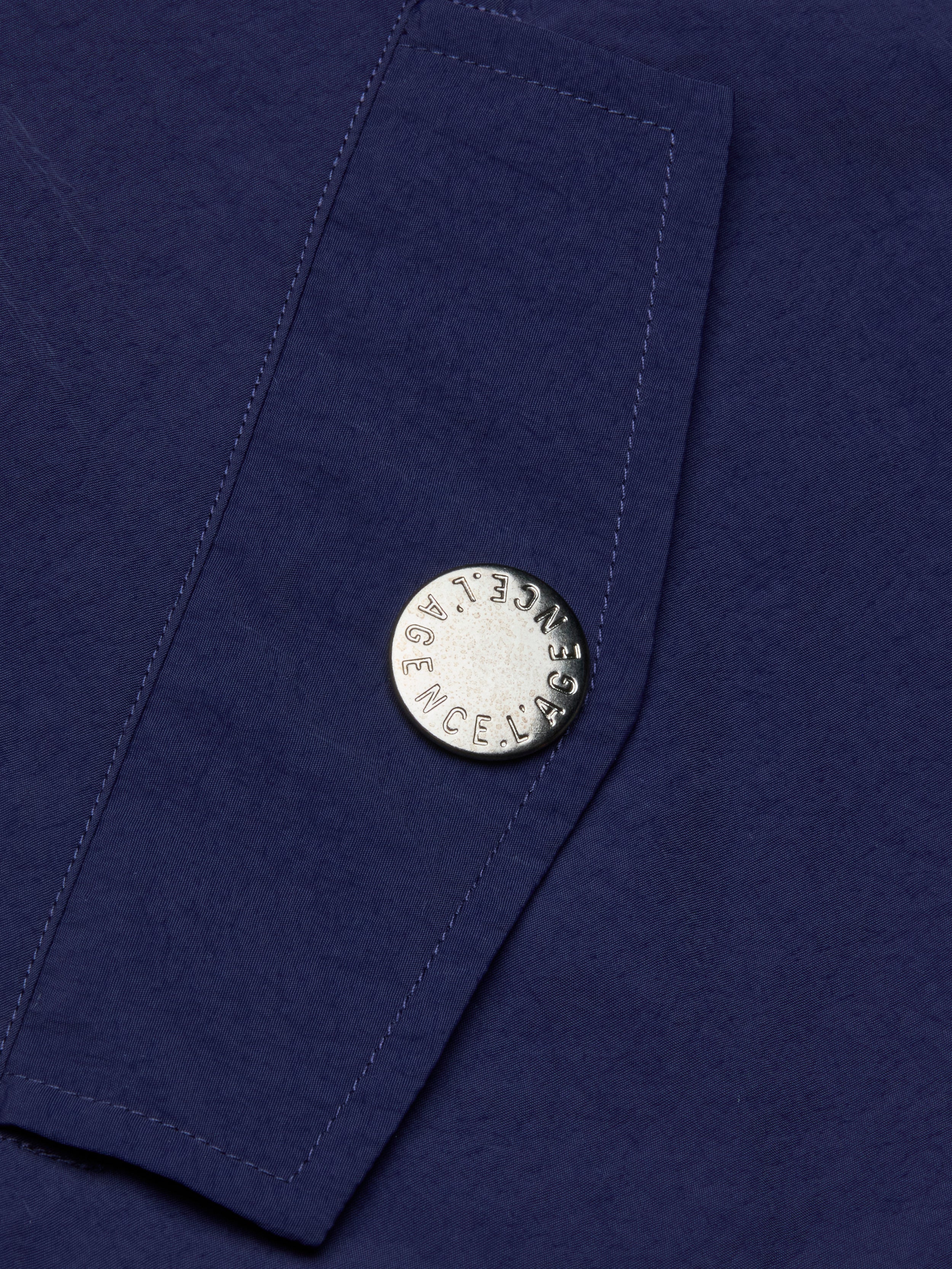 A close-up of the Geovana Hooded Windbreaker’s blue, water-repellent fabric with a silver button stamped LAGENCE. Visible stitching runs diagonally beside the button.