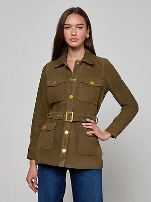 A woman with long, straight brown hair stands against a plain background, wearing the Ishani Suede Jacket—an olive green utilitarian style with gold buttons and four front pockets—paired with blue jeans, looking directly at the camera.