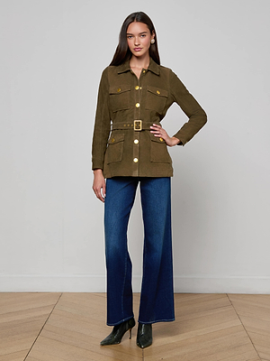 A woman with straight brown hair poses with one hand on her hip, wearing the Ishani Suede Jacket in luxury olive green with gold buttons, wide-leg blue jeans, and pointed dark green heels against a plain background.