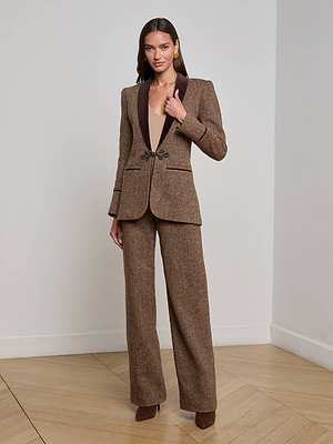 A woman in a minimalist room wears the Gable Herringbone Blazer by L'AGENCE, featuring a velvet shawl lapel and decorative clasp, paired with wide-leg pants—crafted from Italian recycled wool. She has long hair, neutral makeup, and brown heels.