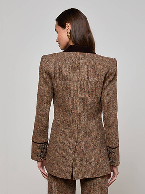 A woman with brown hair models the L'AGENCE Gable Herringbone Blazer, made from Italian recycled wool and featuring embroidered sleeve cuffs, while standing with her back to the camera against a light background.