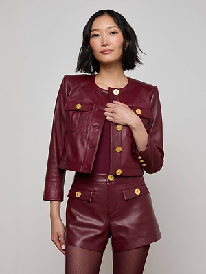 A woman with short black hair wears the Deidra Cropped Leather Jacket in burgundy with gold buttons, matching high-waisted shorts, and a burgundy top, standing against a plain light gray background.