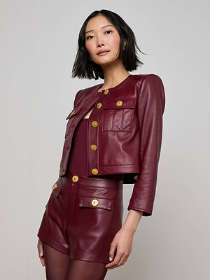 A woman with a bob haircut confidently poses against a plain light background, wearing the Deidra Cropped Leather Jacket in burgundy and matching high-waisted shorts with large gold buttons.
