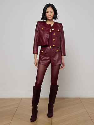 A woman poses indoors against a light-colored wall, wearing the Deidra Cropped Leather Jacket in burgundy Italian lambskin with matching shorts and gold buttons, paired with burgundy tights and knee-high boots on a wooden floor.