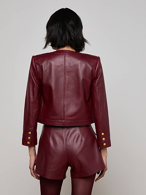 A person with short dark hair is seen from behind wearing the Deidra Cropped Leather Jacket, a collarless Italian lambskin piece with gold sleeve buttons and matching high-waisted shorts. The background is plain and light-colored.