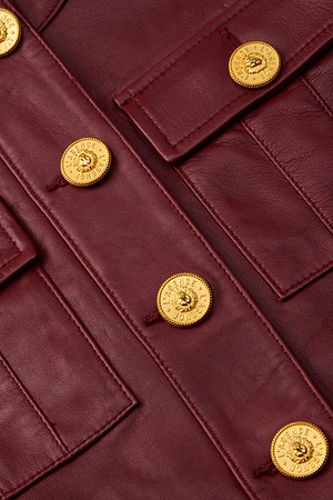 Close-up of the Deidra Cropped Leather Jacket in burgundy Italian lambskin, designed without a collar, with gold embossed buttons, flap pockets, and fine stitching that highlights its luxurious texture.
