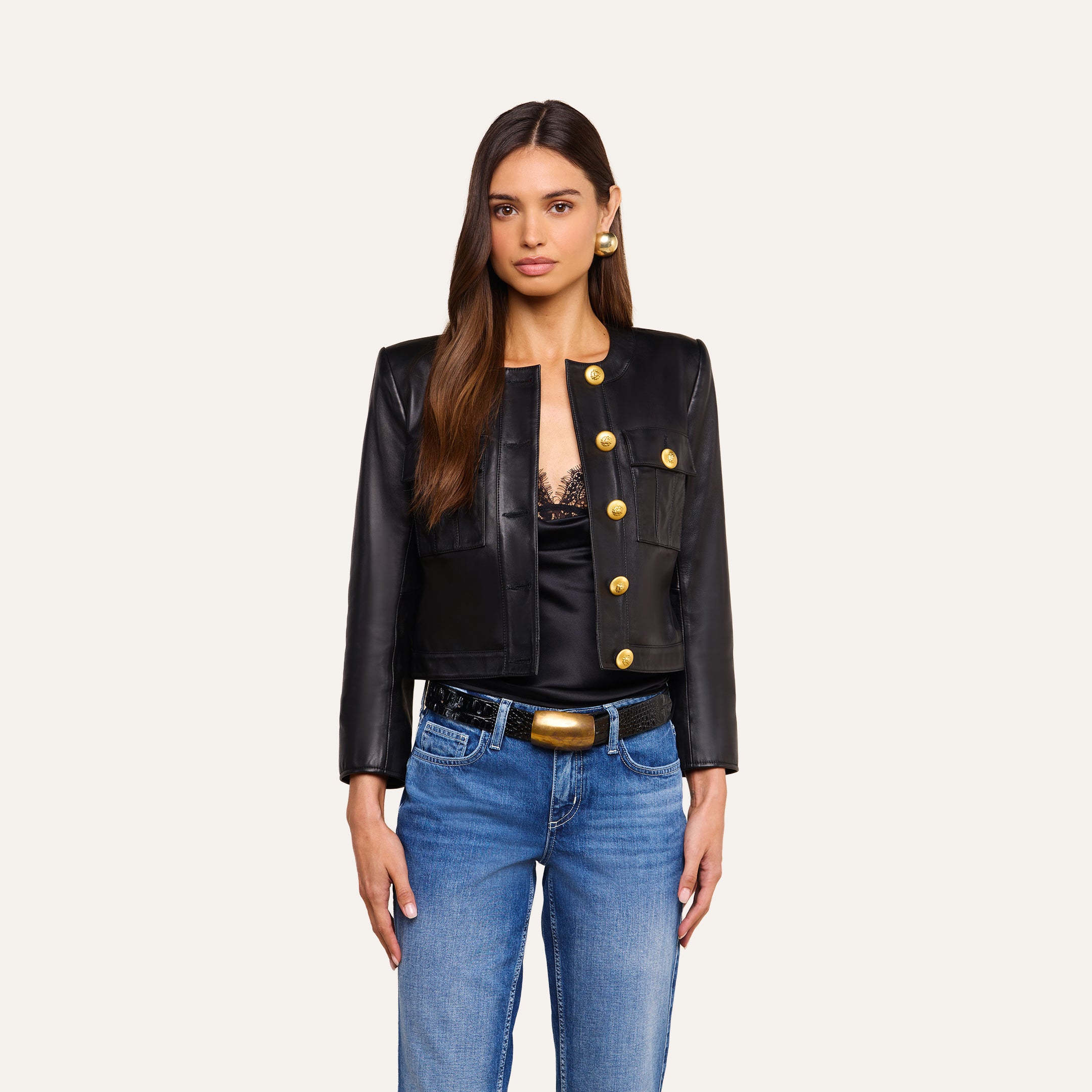 A woman in the Deidra Cropped Leather Jacket—a fitted black leather piece featuring gold accent buttons—pairs it with a black lace top, blue jeans, and gold earrings as she stands facing forward against a plain light background.