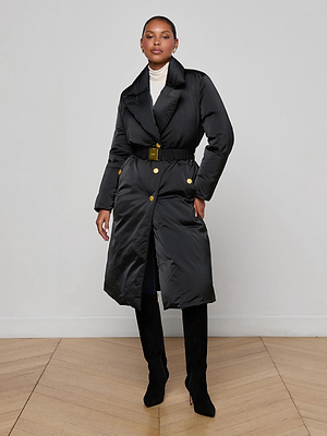 Indoors, a person wears the Kataleya Puffer Coat—a black, water-repellent, belted style with gold buttons—paired with a white turtleneck and black knee-high boots against light walls and a wooden floor.