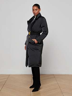A woman stands in a side pose against a light gray wall and wooden floor, wearing the Kataleya Puffer Coat—a long, belted black down coat—paired with black pants and heeled boots.