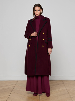 A woman with loose brown hair stands indoors on a wooden floor against a white wall, wearing the Irene Long Coat by L'AGENCE—a maroon women’s double-breasted coat with gold buttons—over a matching turtleneck and wide-legged pants.