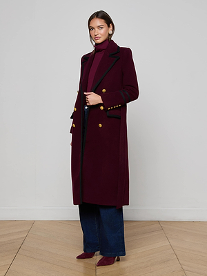A woman poses indoors against a light gray wall and wooden floor, wearing the L’AGENCE Irene Long Coat in burgundy with gold buttons, paired with a matching turtleneck, wide-leg blue jeans, and burgundy heels.