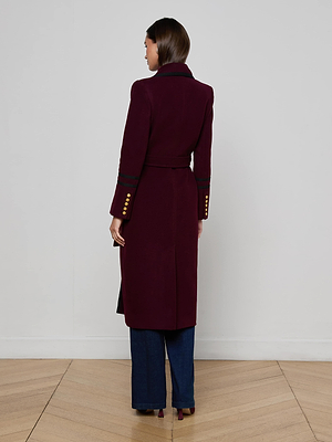 A woman with straight brown hair wears the L'AGENCE Irene Long Coat in maroon, featuring gold cuff buttons, dark sleeve trim, and a belt. She pairs it with dark blue wide-leg pants and heels, facing away from the camera.
