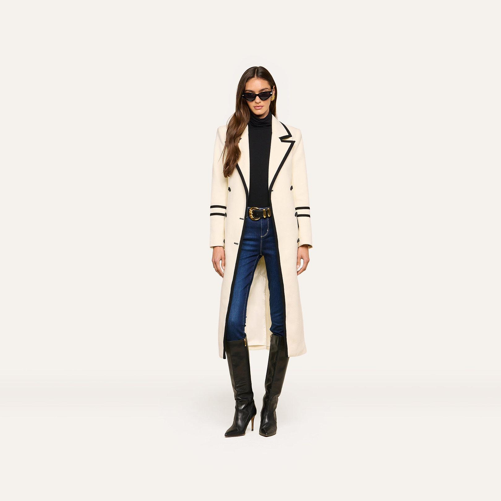 A woman wearing sunglasses, the Irene Long Coat in ivory with black trim, a black turtleneck, blue jeans, and tall black boots stands against a plain white background.
