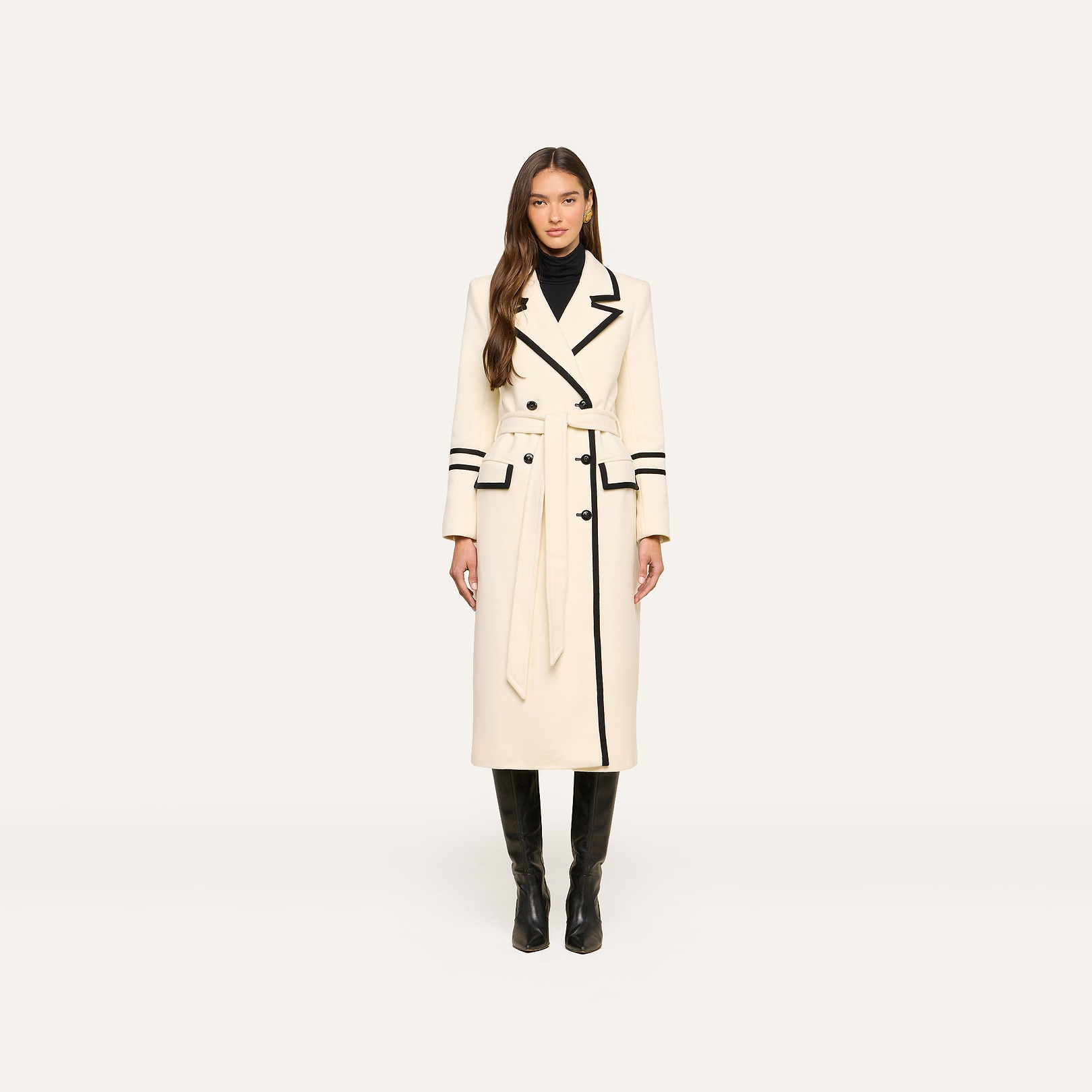 A woman stands facing forward against a plain light background, wearing the Irene Long Coat—a double-breasted long ivory coat with black trim—paired with a black turtleneck and knee-high black boots.