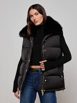 A woman with straight brown hair wears the L'AGENCE Nalo Faux Fur Puffer Vest over a black turtleneck and blue jeans for an effortlessly chic cold-weather look. She stands against a plain light gray background.