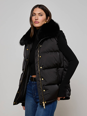 A woman models the L'AGENCE Nalo Faux Fur Puffer Vest—an oversized puffer with a large black faux fur collar—layered over a black long-sleeve top and blue jeans, set against a light gray background.