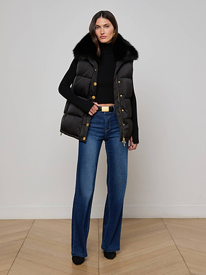 A woman models the L'AGENCE Nalo Faux Fur Puffer Vest with a black turtleneck, blue flared jeans, and black shoes against a white wall—chic cold-weather style inspiration.