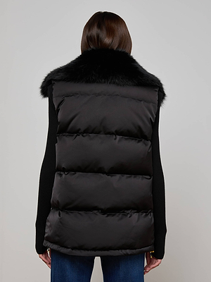 A person with straight brown hair is shown from the back wearing the L'AGENCE Nalo Faux Fur Puffer Vest over a black long-sleeve top and blue jeans, creating a stylish cold-weather look against a plain light background.
