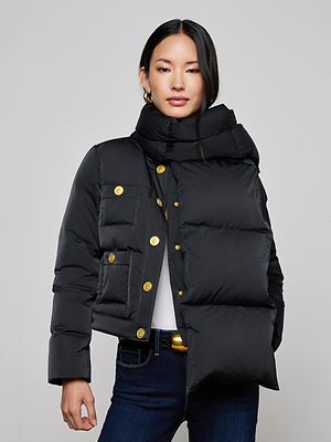 A woman models the Juana Scarf Puffer Jacket, featuring gold buttons, an asymmetric oversized collar, paired with a white top and dark blue jeans, standing against a plain light gray background.